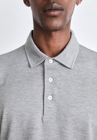Pilkas polo marškinėliai, pagaminti iš tekstūruoto audinio, su apykakle ir trimis baltais mygtukais. Kaklo linija turi sustiprintą apdailą, kad būtų užtikrintas ilgaamžiškumas.