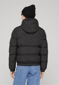 Černá prošívaná bunda s kapucí, s horizontálními prošívanými částmi, elastickými manžetami a krátkým střihem, kombinovaná se světle modrými džíny.