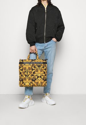 Person i sort quiltet jakke og lyseblå jeans, der holder en sort og guld Versace shopper, iført hvide og guld sneakers.
