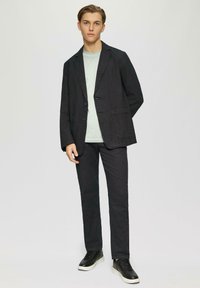 Schwarzer Leinenblazer mit zwei Knöpfen, seitlichen Taschen; hellgrünes Hemd darunter; schwarze Hose; schwarze Sneaker mit weißen Sohlen.