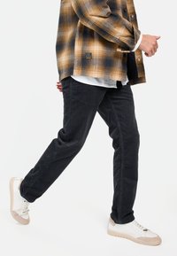 Zwarte corduroybroek met een tapered fit, gecombineerd met een geruite shirt in bruine en marineblauwe tinten, en witte sneakers met beige accenten.