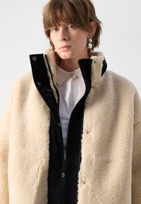Beige schapenleren jas met een hoge kraag, zwarte ritssluiting aan de voorkant en ribbeltextuur accenten. Model draagt een witte shirt eronder.