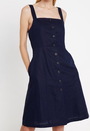Mujer con un vestido azul marino sin mangas con tirantes anchos y botones negros en la parte delantera, con las manos en los bolsillos laterales.