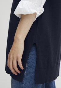 Maglione a maglia blu navy con uno spacco laterale, indossato sopra una camicia bianca e abbinato a jeans. Orlo a coste visibile e texture liscia.