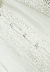 Camicia di tessuto bianco con un design testurizzato, bordi in pizzo e bottoni decorativi. La superficie presenta righe verticali e una finitura liscia.