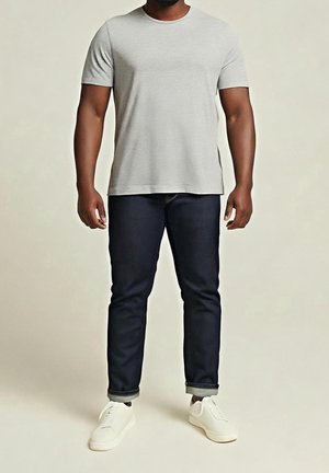 Mann trägt ein hellgraues T-Shirt mit kurzen Ärmeln, dunkelblaue hochgekrempelte Jeans und weiße Turnschuhe, steht vor einem schlichten, hellen Hintergrund.