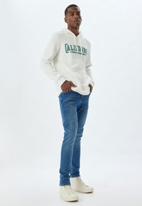 Sudadera blanca con un logotipo verde, bolsillo frontal y cordón. Combinada con jeans azules y zapatillas beige. Textura suave en toda la prenda.