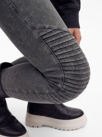Persona inginocchiata che indossa pantaloni grigi con dettagli a coste, giacca nera e stivali neri con suole bianche spesse su sfondo bianco.