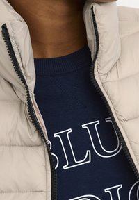 Beige donzen jas met een hoge kraag, open geritst om een donkerblauwe trui met witte letters te laten zien. Gestructureerde stof en contrasterende rits.