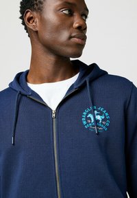 Felpa con zip blu navy in misto cotone, con logo teal e grafica di un cavallo, cappuccio con cordino e dettagli in metallo sulla zip.
