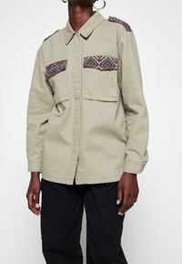 Khaki shirt jacket with two front pockets, patterned fabric accents on pockets and shoulders, long sleeves, and button closure.