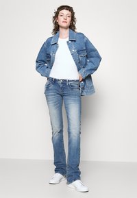 Lys blå denimjakke, hvit t-skjorte, mørkeblå bukser og hvite joggesko. Avslappet passform med klassisk design og lett falmet tekstur.