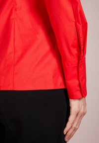 Rode shirt met een gladde textuur, getailleerde pasvorm en knoopmanchetten. Zichtbaar zijnaaddetail. Gecombineerd met een zwart kledingstuk.
