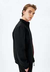 Veste noire zippée avec un col haut, des poignets élastiques et une coupe décontractée. Fabriquée en matériau lisse avec un léger éclat.