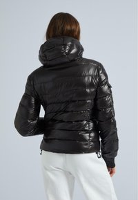 Frau, die eine glänzende schwarze Daunenjacke mit Kapuze trägt, von hinten betrachtet, kombiniert mit weißen Hosen vor einem schlichten Hintergrund.