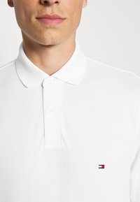Valkoinen polo-paita, jossa on klassinen kaulus, kaksi nappia ja pieni brodeerattu logo vasemmassa rinnassa tummansinisessä, punaisessa ja valkoisessa. Tekstuuroitu kangas.