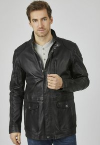 Giubbotto di pelle nera con colletto alto, chiusura con zip e bottoni a pressione, tasche frontali e una texture liscia. Il modello tiene il giubbotto leggermente aperto.