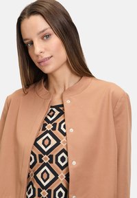 Braune leichtejacke mit runder Halslinie und Druckknöpfen, über einem gemusterten Kleid mit schwarzen, weißen und beigen geometrischen Formen.