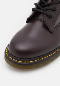 Acercamiento a una bota de cuero marrón oscuro con cordones negros, costura amarilla alrededor de la suela y suela de goma translúcida sobre un fondo blanco.