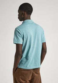 Pikétröja i ljusbl ått, kortärmad, med texturerad tyg. Bakvy visar kragen och en avslappnad passform, tillsammans med bruna byxor.