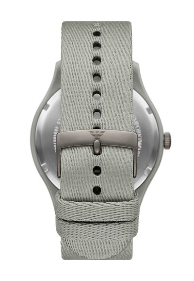 Bracelet de montre en tissu gris clair avec des trous ajustables, doté d'une boucle métallique avec logo et d'un cadran de montre métallique visible en arrière-plan.