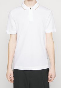 Vit polo-skjorta med texturerad tyg, korta ärmar och krage med guldaccenter. Har en knappstängning och subtil logodetalj.