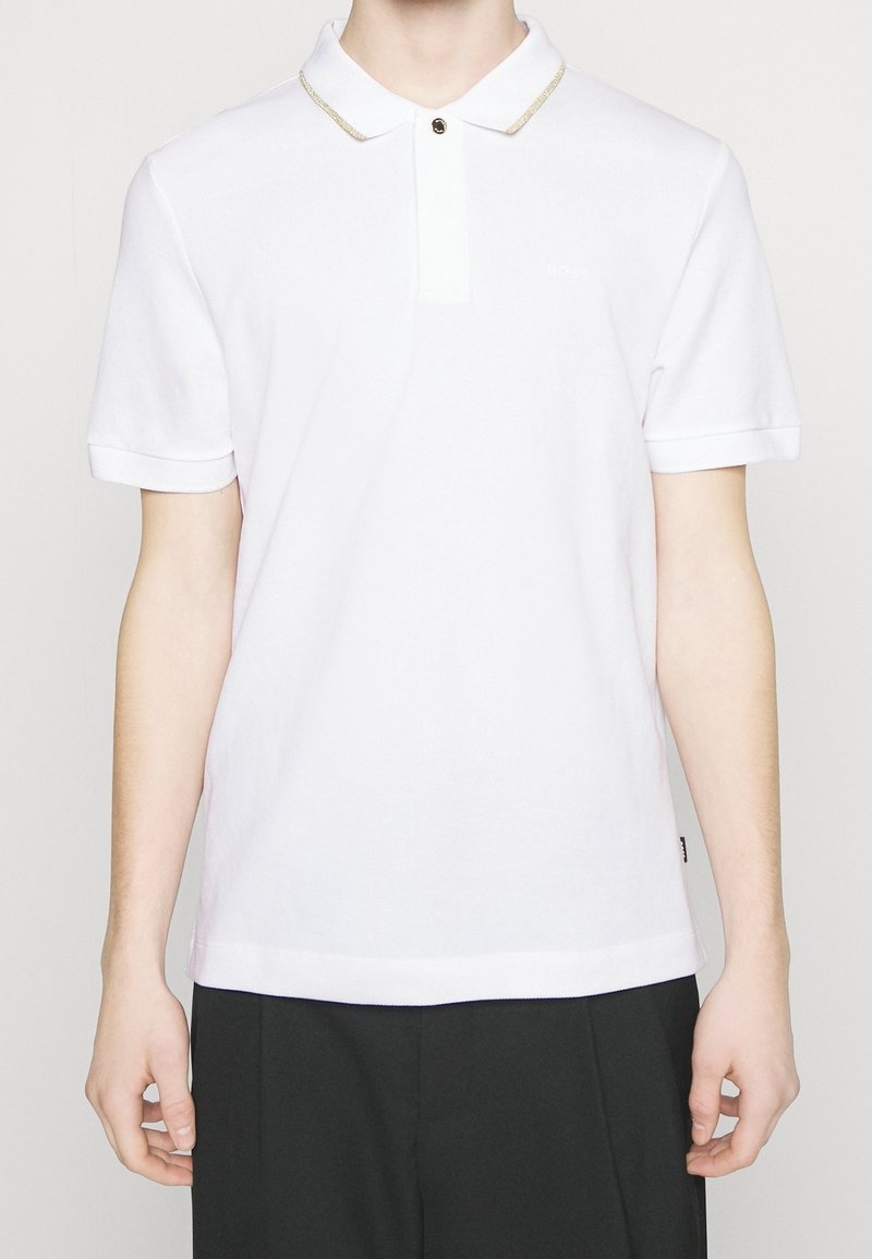 Vit polo-skjorta med texturerad tyg, korta ärmar och krage med guldaccenter. Har en knappstängning och subtil logodetalj.