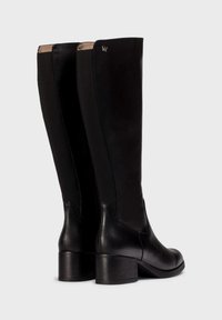 Botas de cuero y tela negras hasta la rodilla con un tacón cuadrado. Presentan una textura suave y un discreto detalle de logo cerca de la parte superior.