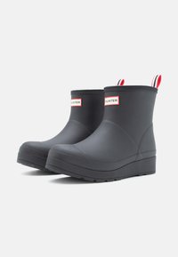 Bottes de cheville en caoutchouc noires avec un bout arrondi, dotées d'une semelle compensée, d'une languette en rouge et blanc à rayures, et du logo "HUNTER" sur le côté.