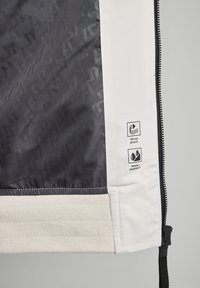 Giacca antivento e idrorepellente nera e grigia con orlo bianco a coste; presenta simboli di cura sul lato, che mostrano il materiale e la consistenza.