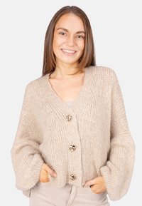Beige gestrickte Strickjacke mit V-Ausschnitt, übergroßen Ärmeln und drei dekorativen Knöpfen. Weiche Textur und grob gestricktes Design.