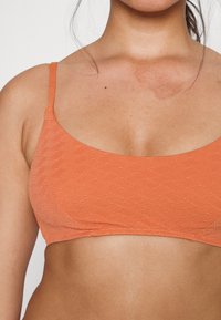 Bralette laranja texturizado com alças finas, apresentando um padrão geométrico e um decote redondo, enfatizando um design suave e ajustado.