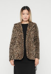 Žena oblečená v hnedo-čiernej leopardej vzorovanej priestrannej bunde s dlhým rukávom čiernej šaty stojí pred jednoduchým bielym pozadím.