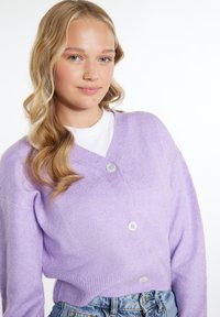 Fialový cardigan s výstřihem do V, oversized střihu, se třemi knoflíky vpředu a žebrovaným spodním lemem. Nosený přes bílé tričko a džínové šortky.