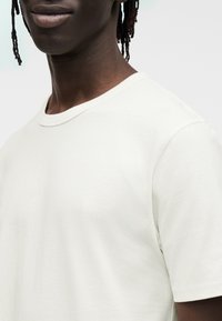 Maglietta in cotone color crema con scollo rotondo, maniche corte e tessitura liscia, mostrata dal petto in su su uno sfondo neutro.