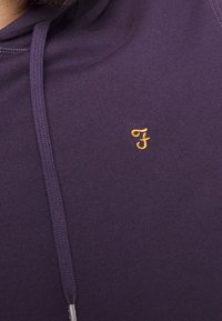 Felpa di cotone viola con cappuccio, caratterizzata da un cordino e un piccolo logo ricamato in oro. Il tessuto ha una consistenza morbida e una finitura liscia.