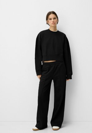 Felpa nera corta abbinata a pantaloni neri a vestibilità rilassata. Entrambi sono realizzati in un tessuto morbido, presentano un design semplice e una cintura elastica.