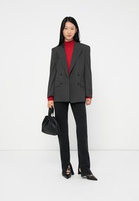 Woman wearing a gray pinstripe oversized blazer, red turtleneck, black pants with front slits, black pointed shoes, holding a black handbag.