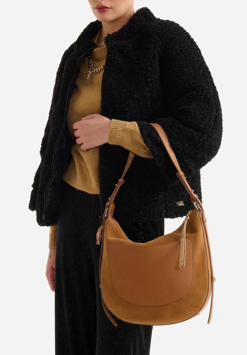 Borsa a mano in pelle e camoscio marrone con forma curva, tasca sporgente e dettagli in metallo dorato, tenuta da una persona vestita di nero e oro.