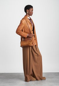 Ruskea nahkableiseri mustilla nappeilla, yhdistetty ylisuurien ruskeiden housujen kanssa. Pieni ruskea laukku, jossa on hihna ja kultaiset metalliosat, pidetään kädessä.