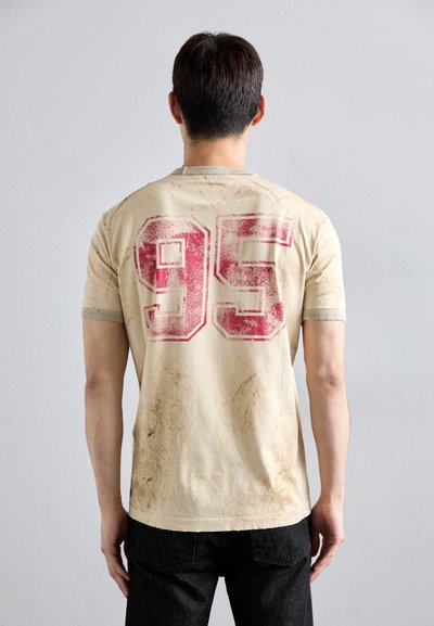 Hellbeige Baumwoll-T-Shirt mit einer großen roten "95" auf dem Rücken, sichtbaren Schmutzflecken und einem klassischen Rundhalsausschnitt.