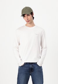 Witte katoenen T-shirt met lange mouwen en een klein logo op de linkerborst, gecombineerd met blauwe denim jeans en een olijfgroene baseballpet.