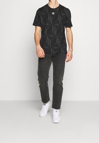 Hombre vestido con una camiseta negra de Adidas con estampado, jeans gris oscuro y zapatillas blancas de Adidas, de pie contra un fondo claro y uniforme.