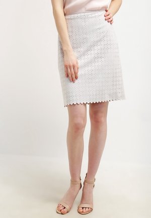 Frau trägt einen weißen, perforierten A-Linien-Rock mit wellenförmigem Saum und nudefarbenen Sandalen mit Knöchelriemen, steht vor einem einfarbigen Hintergrund.