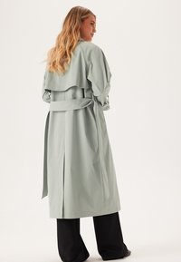 Ljusgrön trenchcoat med två sidofickor, bälte i midjan och en lagerkrage-design, tillverkad av ett slätt, lätt tyg.
