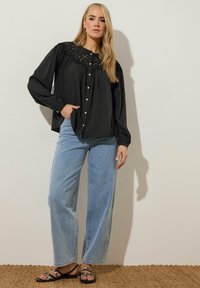 Svart knäppt skjorta med en texturerad yoke, långa ärmar och en avslappnad passform, ihop med ljusblå vida jeans och svarta sandaletter.