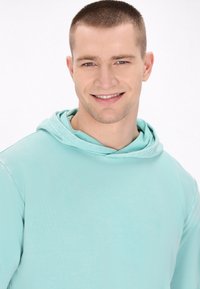 Világoskék kapucnis pulóver, puha tapintással, elülső zsebbel és húzózsinóros kapucnival. Az anyag simának és rugalmasnak tűnik.