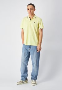 Żółta bawełniana koszulka polo z czarnymi akcentami przy kołnierzyku, w połączeniu z jasnoniebieskimi dżinsami i beżowymi sneakersami z zielonymi i białymi detalami.