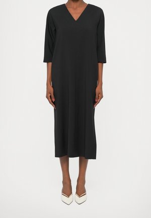 Femme portant une robe midi noire à manches trois-quarts et des talons blancs pointus, se tenant devant un fond blanc uni.