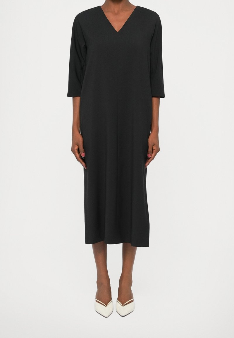 Femme portant une robe midi noire à manches trois-quarts et des talons blancs pointus, se tenant devant un fond blanc uni.